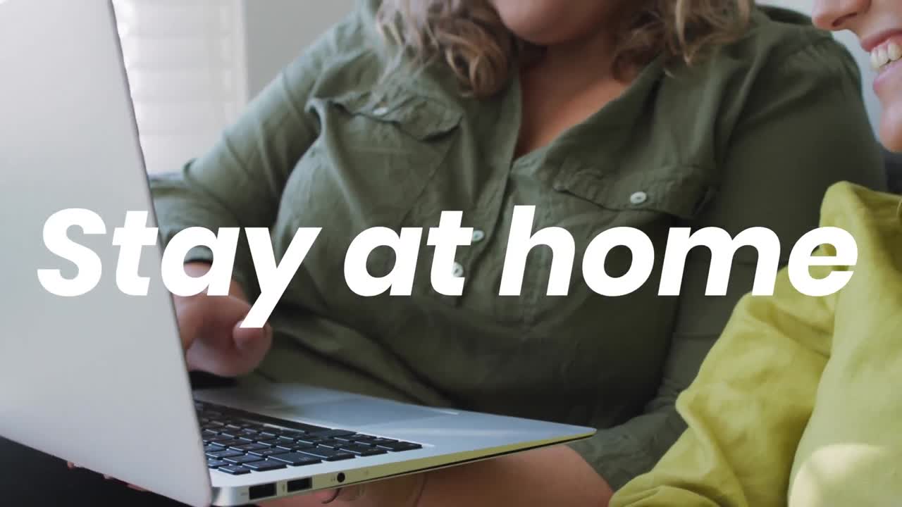 Animation of stay at home text over caucasian female couple using laptop
