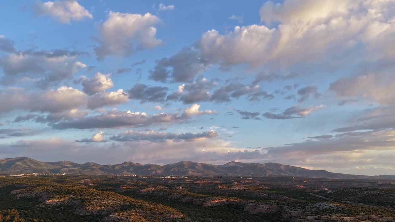 Timelapse con drone 4K UHD: il vasto cielo mattutino domina la complessa topografia di una mesa di macchia. La luce dell'alba proietta profonde ombre strutturali sul terreno e illumina nuvole sparse color pastello.