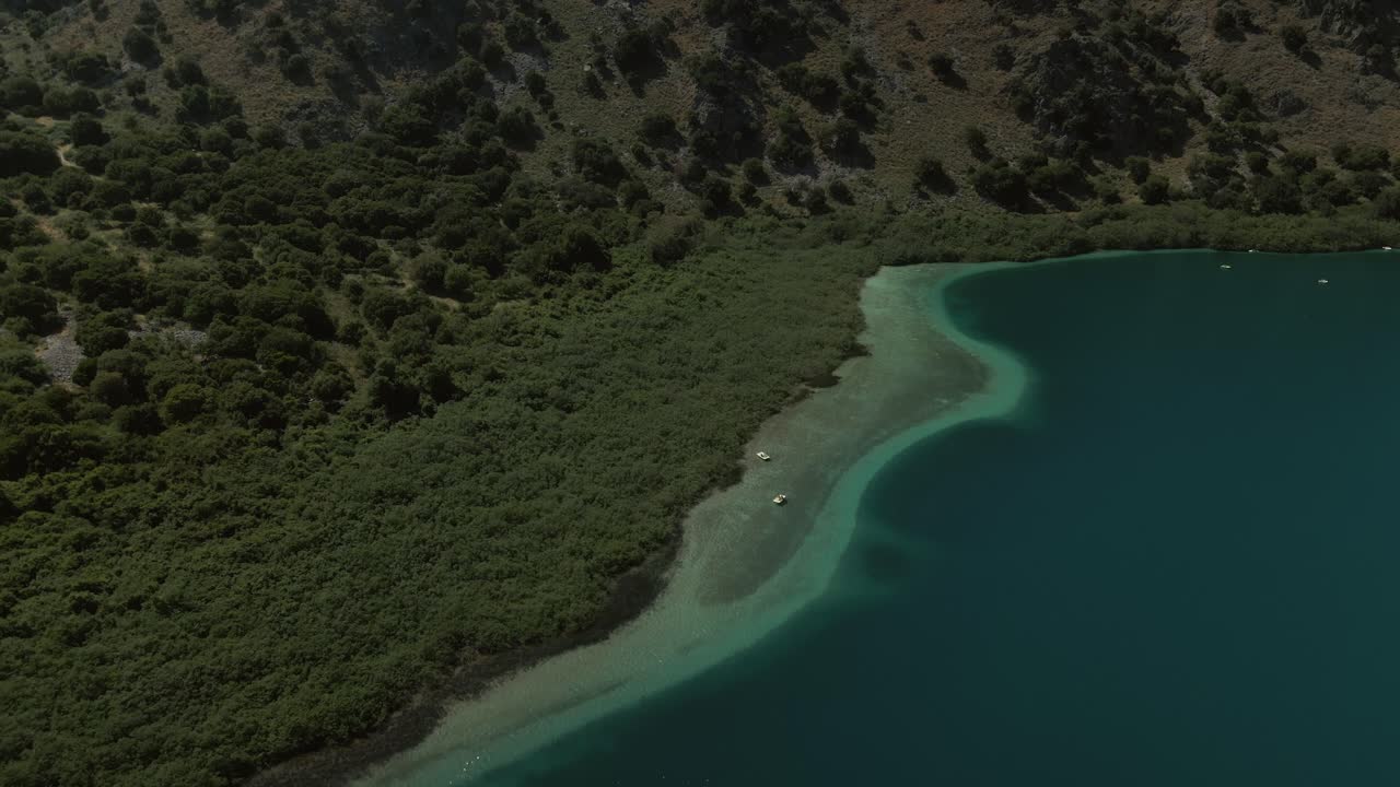 Small boats rest on the turquoise shallows of Lake Kournas in Crete with surrounding lush green vegetation and rugged hillside in the background
