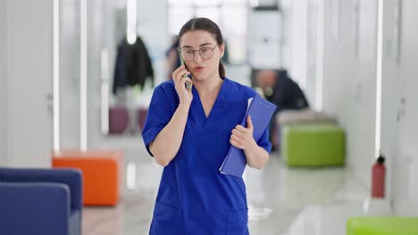 A-confident-brunette-doctor-girl-in-round-glasses-and-a-blue-uniform-walks-along-the-corridor-and-talks-on-the-phone-in-a-modern-clinic.-Confident-brunette-girl-in-round-glasses,-doctor-communicates-goes-on-call-in-a-modern-clinic