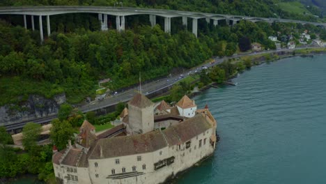 Das-Schloss-Chillon-Liegt-Direkt-Am-Wasser,-Dahinter-Verläuft-Eine-Straße,-Die-Von-Großen-Bäumen-Umgeben-Ist-Und-Auf-Der-Die-Straße-Von-Vielen-Vier-Rädern-Und-Vielen-Fahrzeugen-Befahren-Wird