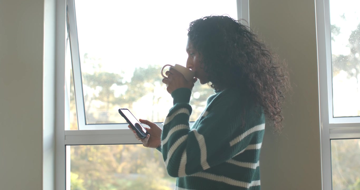 Donna afroamericana vicino alla finestra, che alterna il tenere il telefono e il sorseggiare da una tazza, con un maglione verde
