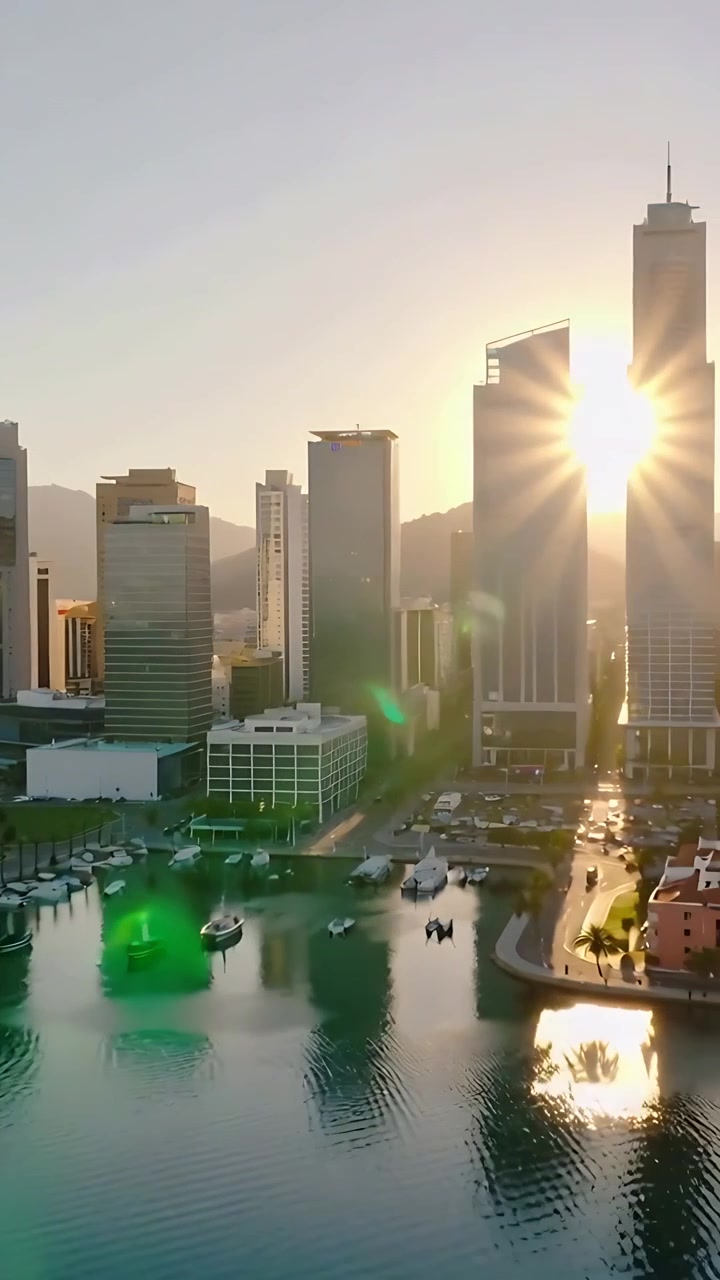 Vídeo vertical: Drone em panorâmica revelando torres sobre o porto, com reflexos do sol atingindo a água e os barcos.