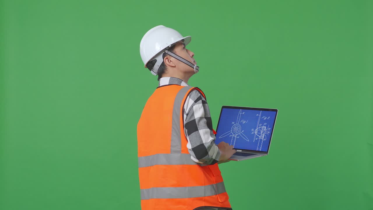 Back View Of Asian Male Engineer With Safety Helmet Looking At The Wind Turbine Blueprint On Laptop While Standing In The Green Screen Background Studio