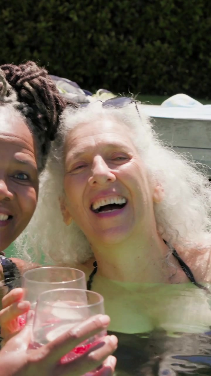 Verticale video: Volwassen vrouw draait zich om naar een vriendin die roept, met een roze drankje, lacht en nipt in het zwembad.
