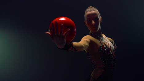 chica graciosa rodando la pelota en las manos en el interior. gimnasta haciendo ejercicio en el espacio oscuro.