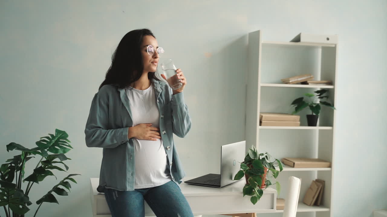 Young pregnant woman massaging her belly and drinking water at home