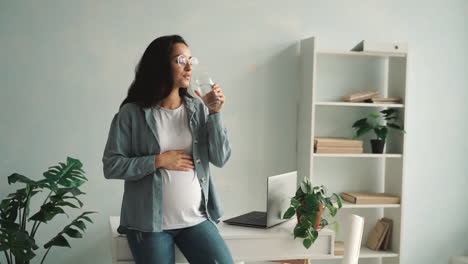 Young-pregnant-woman-massaging-her-belly-and-drinking-water-at-home