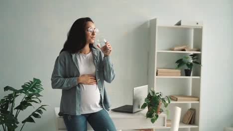 young pregnant woman massaging her belly and drinking water at home