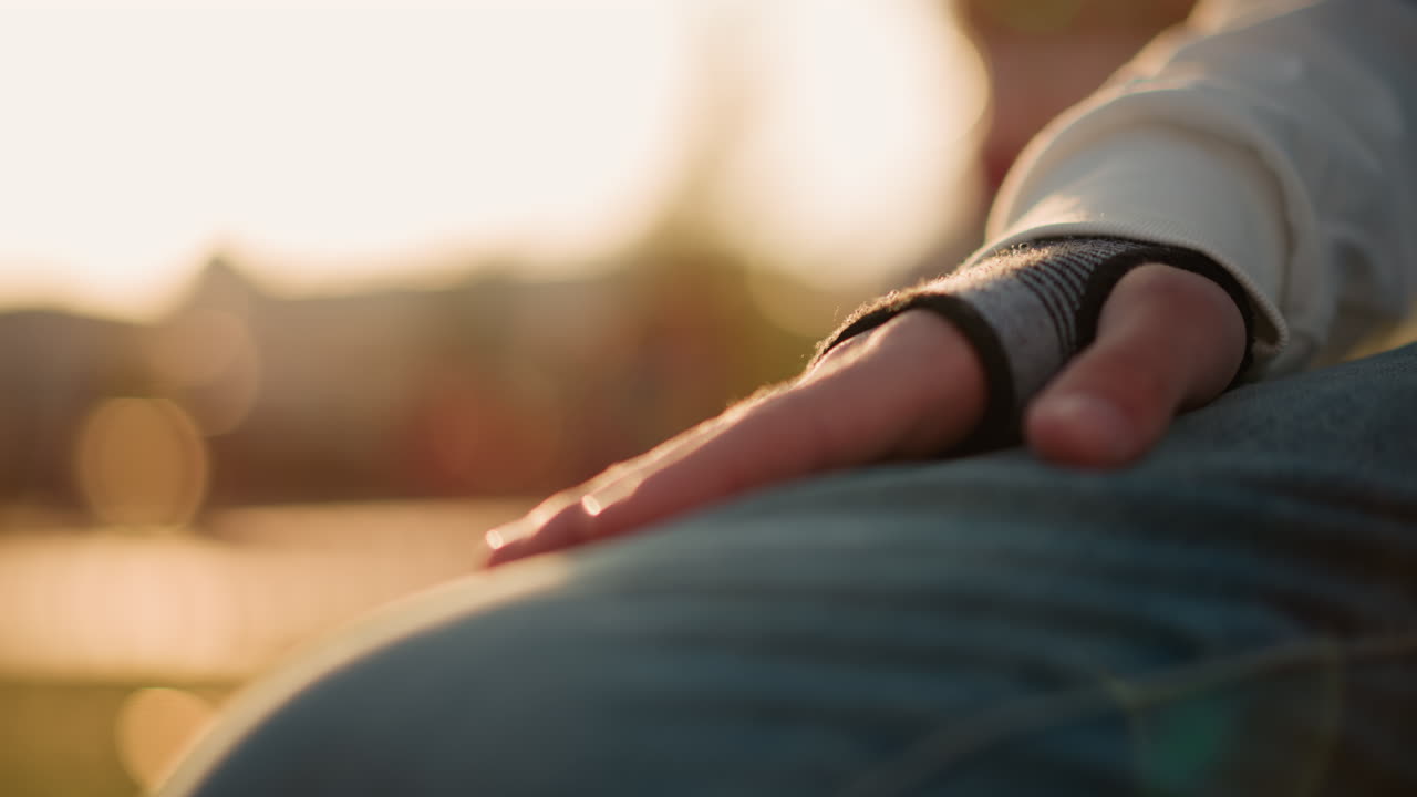 primo piano di una mano appoggiata su un ginocchio, il bagliore del tramonto scalda il denim testurizzato e il guanto senza dita, un momento intimo di un musicista che evoca calma e quieta preparazione prima di provare in un parco del quartiere