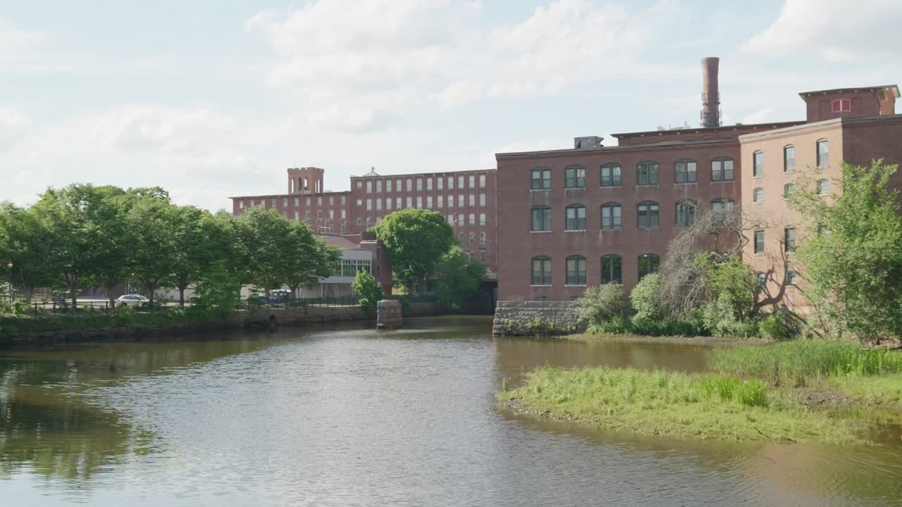 Langsamer Schwenk nach links, der ein historisches Backsteinmühlengebäude an einem Fluss mit Flussgras zeigt und dann Bäume und einen schwarzen Metallzaun entlang eines Gehwegs enthüllt