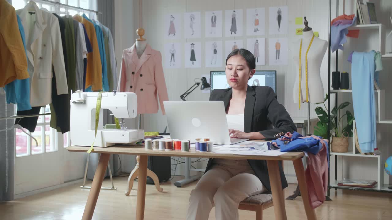 비즈니스 슈트 를 입은 아시아 여성 디자이너 가 노트북 을 사용 하고 스튜디오 에서 옷 을 디자인 하는 동안 스트레칭 을 하고 있다