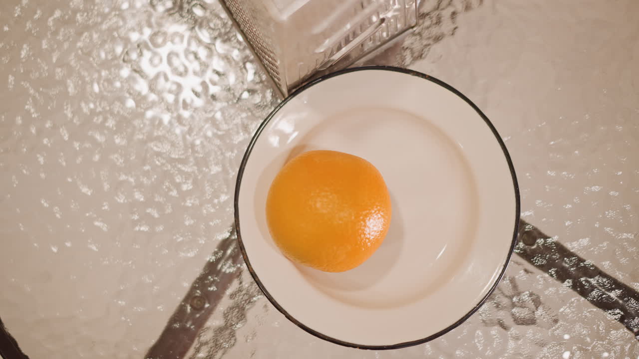 Overhead view orange on white plate. Topdown composition shows single orange resting on enamel rim plate on textured glass table, bright reflection and glossy peel, minimal studio lighting, evokes