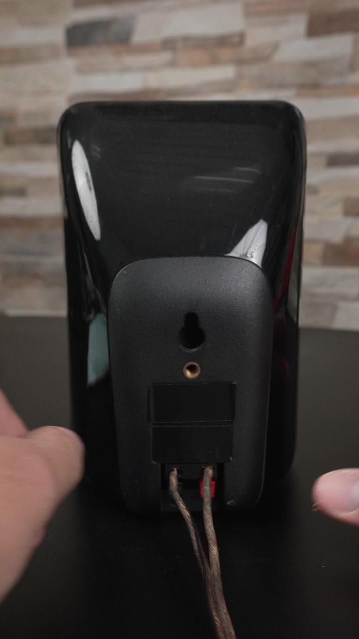 Hands attach bare speaker wire to the spring clip terminals on the back of a black bookshelf speaker on a desk, useful for tutorials, product demos, or home o guides.