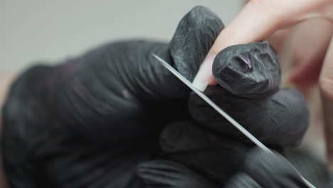 close up image of nail technician wearing black gloves using thin metal nail file to shape customer's fingernail with precision and care during detailed manicure process in clean professional salon