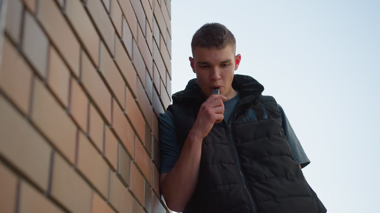 young boy leaning against brick wall outdoors inhaling shisha smoke into mouth as he coughs looking at vape device wearing black puffer vest over dark shirt in sunlight casting shadows on bricks
