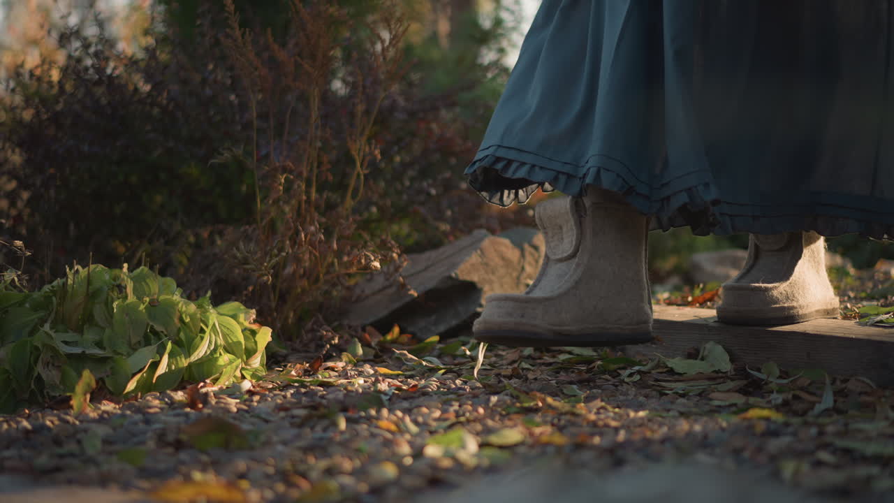 Woman walking garden path in boots, evening golden light and long skirt brushing foliage, stepping over stone slabs and scattered leaves, home gardener foraging lettuce and herbs, deliberate slow