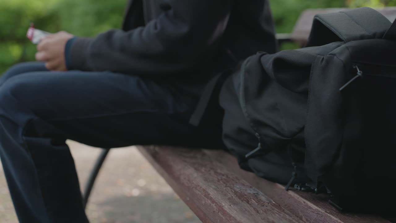 wet park bench with black backpack, man sits in dark hoodie and jeans, hands resting on knees, damp wooden slats and metal armrest, suburban greenery blurred in background, tense quiet atmosphere