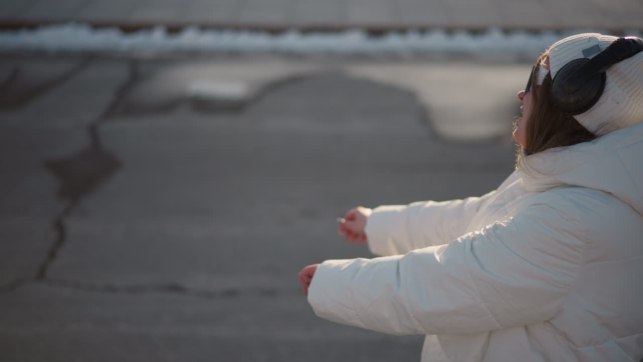 Young girl, dressed in white winter jacket and beanie, moves carefully while enjoying rhythm in outdoor setting, her cautious gestures and smiling expression shine under soft daylight