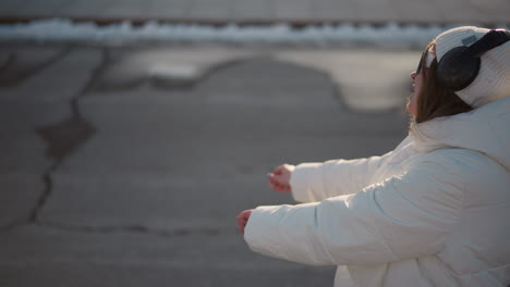 young girl, dressed in white winter jacket and beanie, moves carefully while enjoying rhythm in outdoor setting, her cautious gestures and smiling expression shine under soft daylight