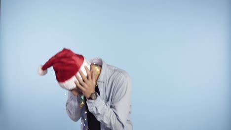 young crazy guy in traditional santa hat shaking, waving head head, getting crazy - holding hands on face, grimace while standing isolated over blue background