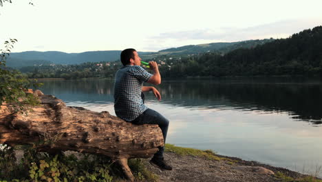 a man sits alone on a tree stump looking out over a lake as he drinks a beer