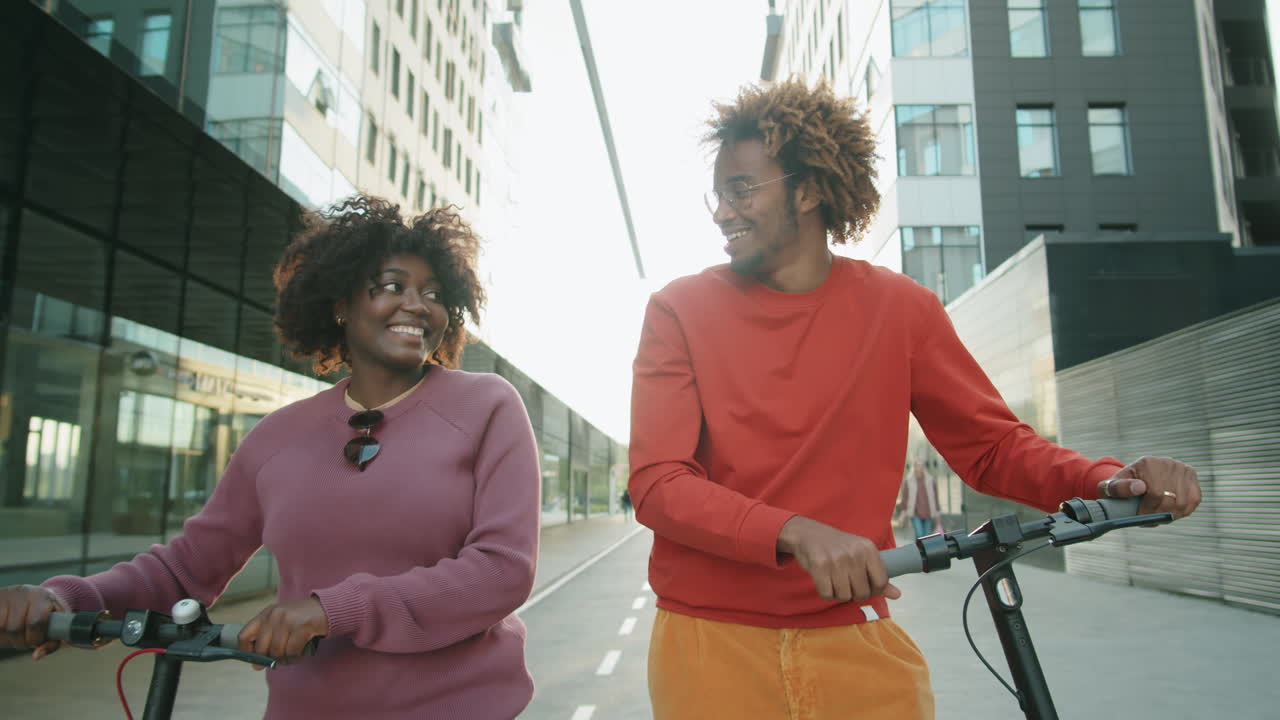 amigos afroamericanos caminando con scooters eléctricos en la ciudad y hablando