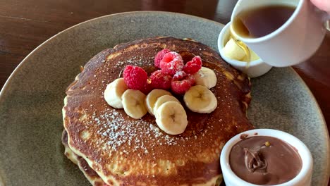 Verter-Jarabe-A-Mano-Sobre-Una-Pila-De-Tortitas-De-Suero-De-Leche-Con-Frambuesas-Frescas-Y-Rodajas-De-Plátano-Encima