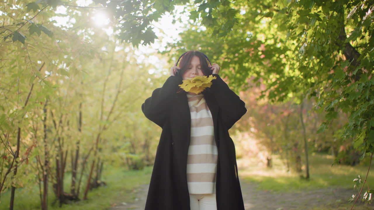 Nature walker standing with bunch of leaves in mouth while adjusting headphones, moving body with excitement under bright sunlight surrounded by lush greenery and autumn atmosphere in forest pathway