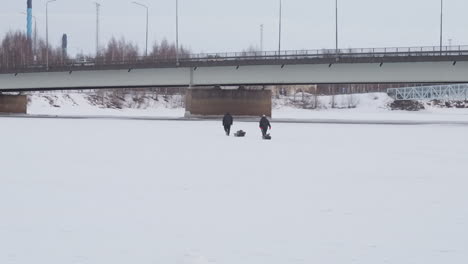 Two Fishermen Walk on Frozen River in Wintertime, Cars Crossing Bridge