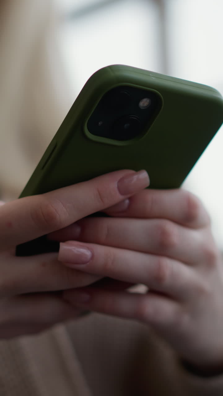 primer plano de una mujer sosteniendo un iphone y operándolo, con enfoque suave en el teléfono y las manos, ventana de vidrio visible en el fondo, enfoque en la interacción del dispositivo móvil en un entorno moderno