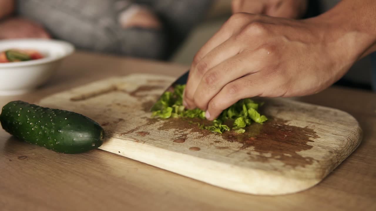 Close up slow motion footage of a young mulatto guy cutting lettuce for salad, making lunch with his girlfriend who's sitting beside him on the kitchen table surface.