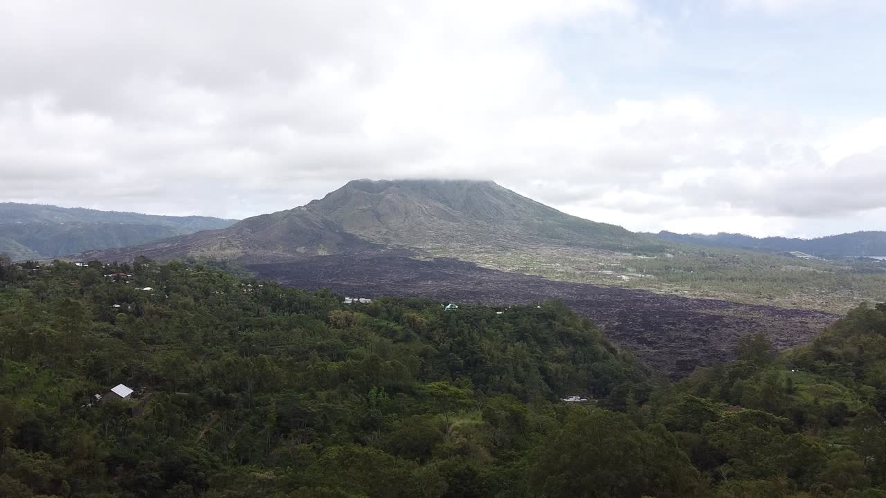 Ripresa aerea con drone di un paesaggio montano vulcanico a Bali, in Indonesia, che mostra colline boscose, campi di lava scura e un cielo nuvoloso che creano uno scenario naturale spettacolare