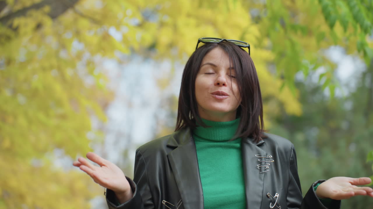 Donna nel parco autunnale con giacca di pelle, dolcevita verde, occhiali da sole appoggiati sulla testa, gesti espressivi con le mani mentre parla alla fotocamera, sfondo con caldo fogliame giallo, atmosfera rilassata e sicura di sé