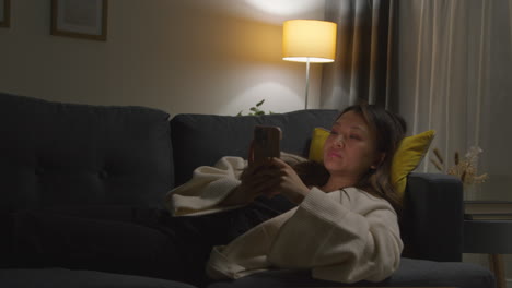 woman spending evening at home lying on sofa with mobile phone scrolling through internet or social media