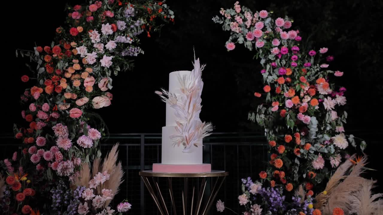 Elegant white wedding cake decorated with feathers and flowers, displayed on a table under a floral arch at night
