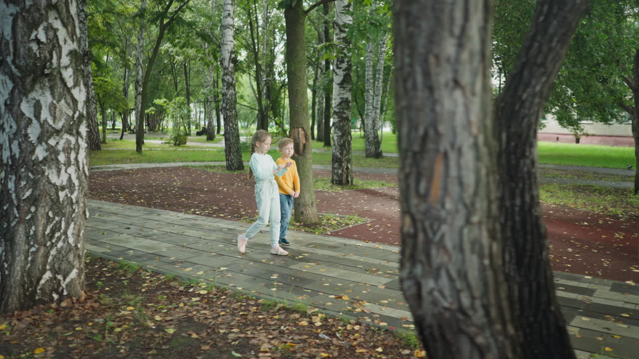 Boy and girl walk side by side on park pathway surrounded by birch trees, dry leaves, and green grass, sharing friendly interaction in calm outdoor setting with soft daylight after rainfall