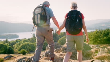 Slow-Motion-Rear-View-Shot-Of-Senior-Couple-Standing-At-Top-Of-Hill-Looking-At-Beautiful-Countryside-On-Hike-Through-Lake-District-In-UK