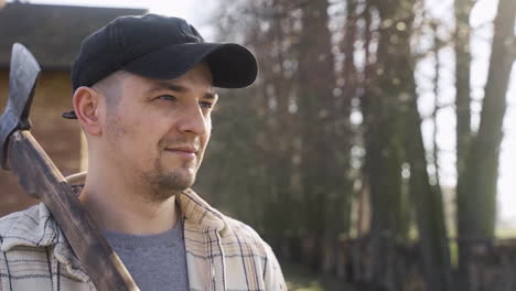 close-up view of caucasian man holding an ax outside a country house while smiling and looking at camera