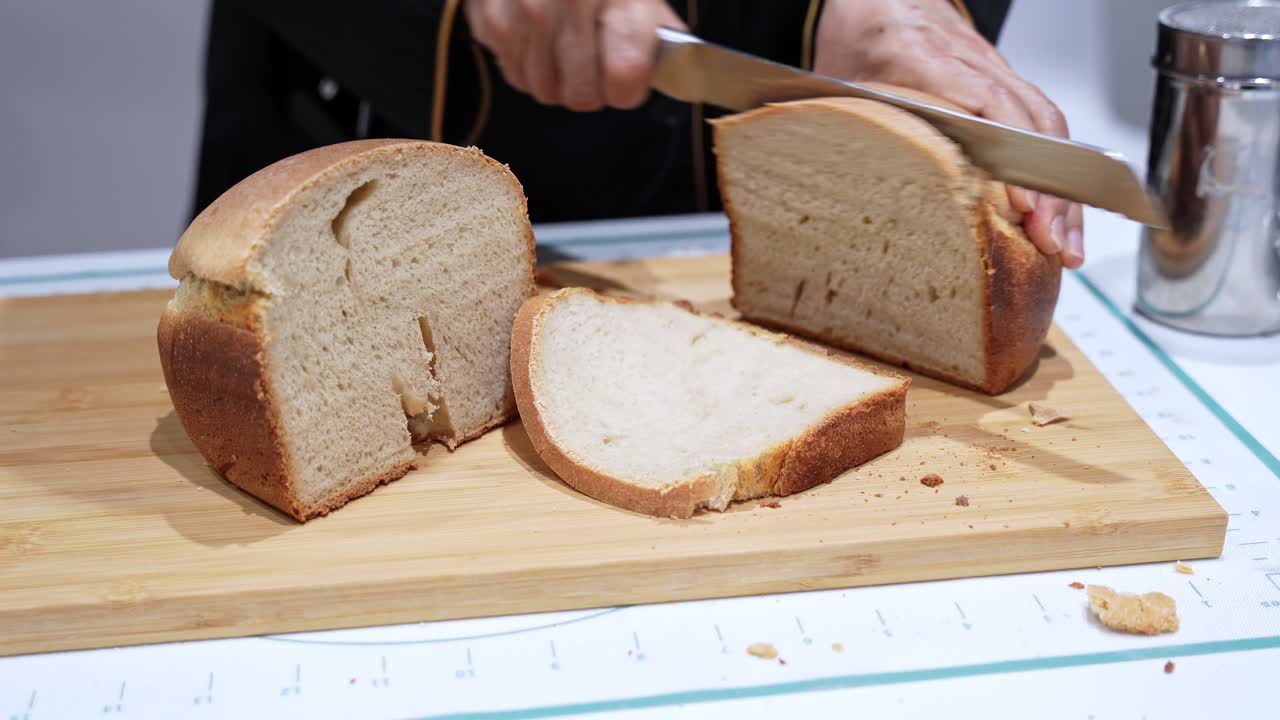 Close up of freshly baked homemade bread loaf being sliced with a knife on a wooden cutting board, showing soft interior texture and golden crust in a kitchen setting