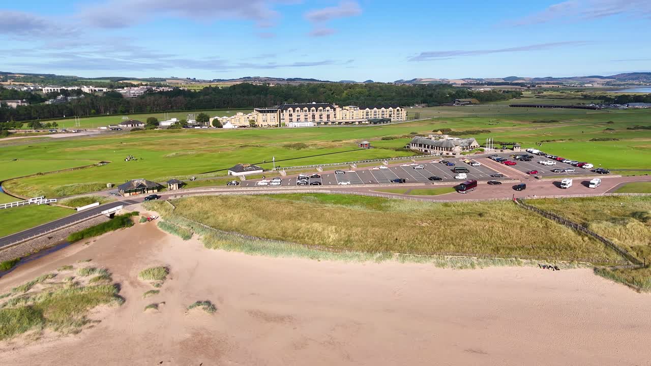 Drone footage glides above sandy beach, grassy dunes, Victorian architecture, and iconic golf course in St Andrews, Scotland, under bright daylight