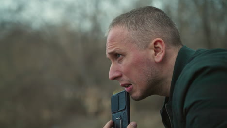 a close view of a man looking intensely to the side, holding a black smartphone near his mouth, the background features a forest