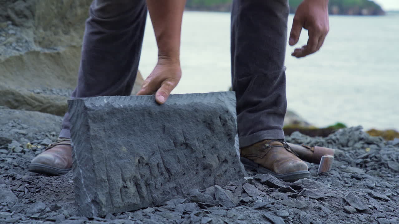 Handheld closeup shot of a cancagua stone craftsman measuring and chipping a large stone slab on the coast line of Ancud, Chiloe Island.