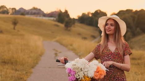 middle plan a young sexy blonde on a bike in a dress and flowers in a basket on the handlebar goes across the field in the summer walking on the grass in slow motion.