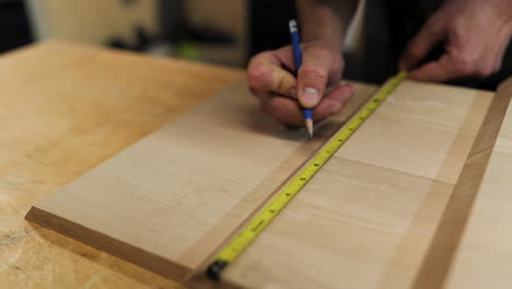 A person measures and marks a piece of wood using a tape measure and pencil, demonstrating attention to detail in a woodworking project in a workshop.