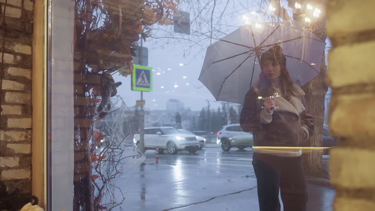 Female holding umbrella walking past cafe, looking through glass window on cold overcast day, wearing black knit cap, brown shearling jacket, black trousers, with passing cars and wet street