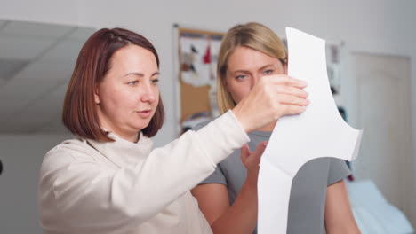 Two female dressmakers examining cloth pattern design closely, smiling and talking while standing in workshop with inspiration boards in background, sharing ideas during creative discussion