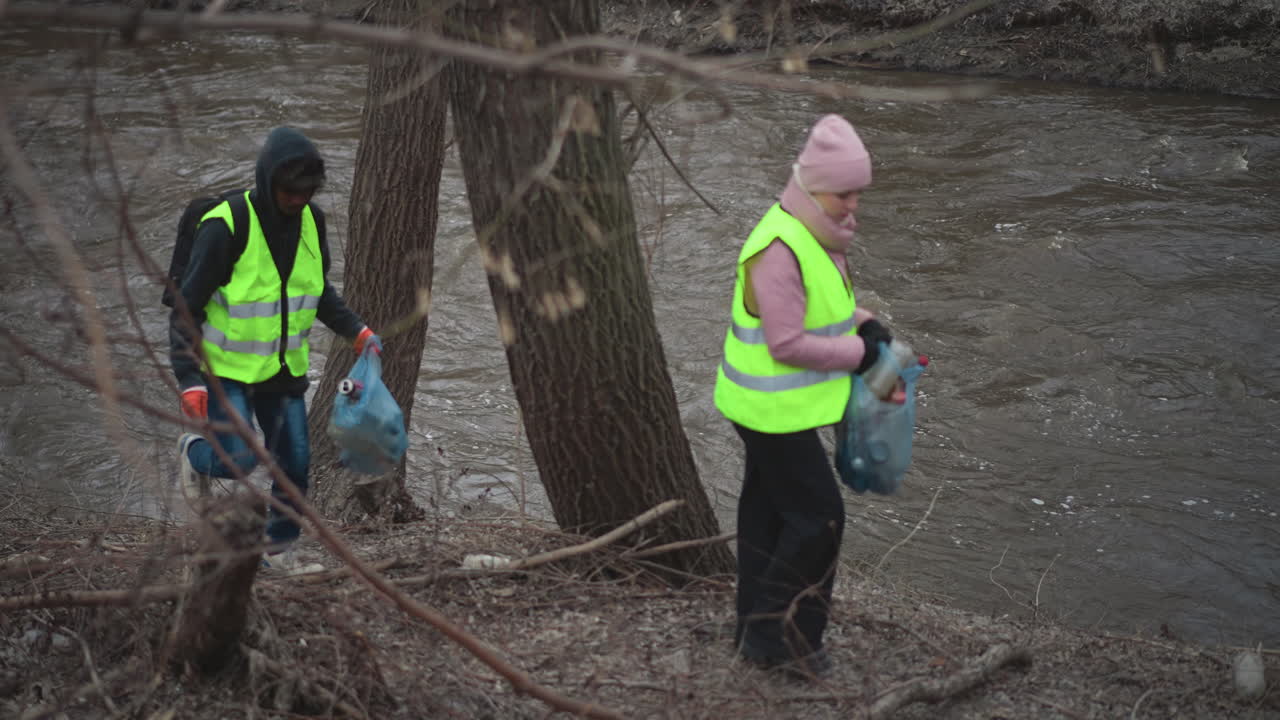 Woman in pink hat and padded jacket with reflective safety vest collects trash in bag along riverbank, holding onto tree for support, man in hooded sweatshirt with safety vest visible in background