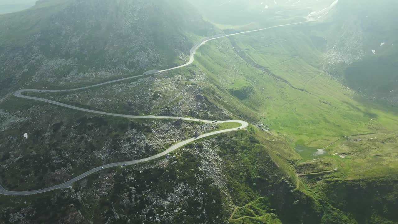 An aerial video captures a winding mountain road snaking through a rugged green landscape. The curves of the road contrast beautifully with the rocky terrain and rolling hills, bathed in soft sunlight