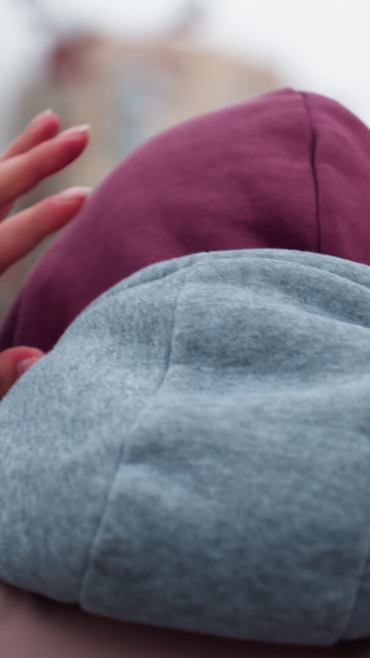 vista de cerca de atrás de un individuo ajustando su gorra mientras camina en la fría temporada de invierno, manos agarrando una capucha cálida, fondo borroso de árboles y edificios